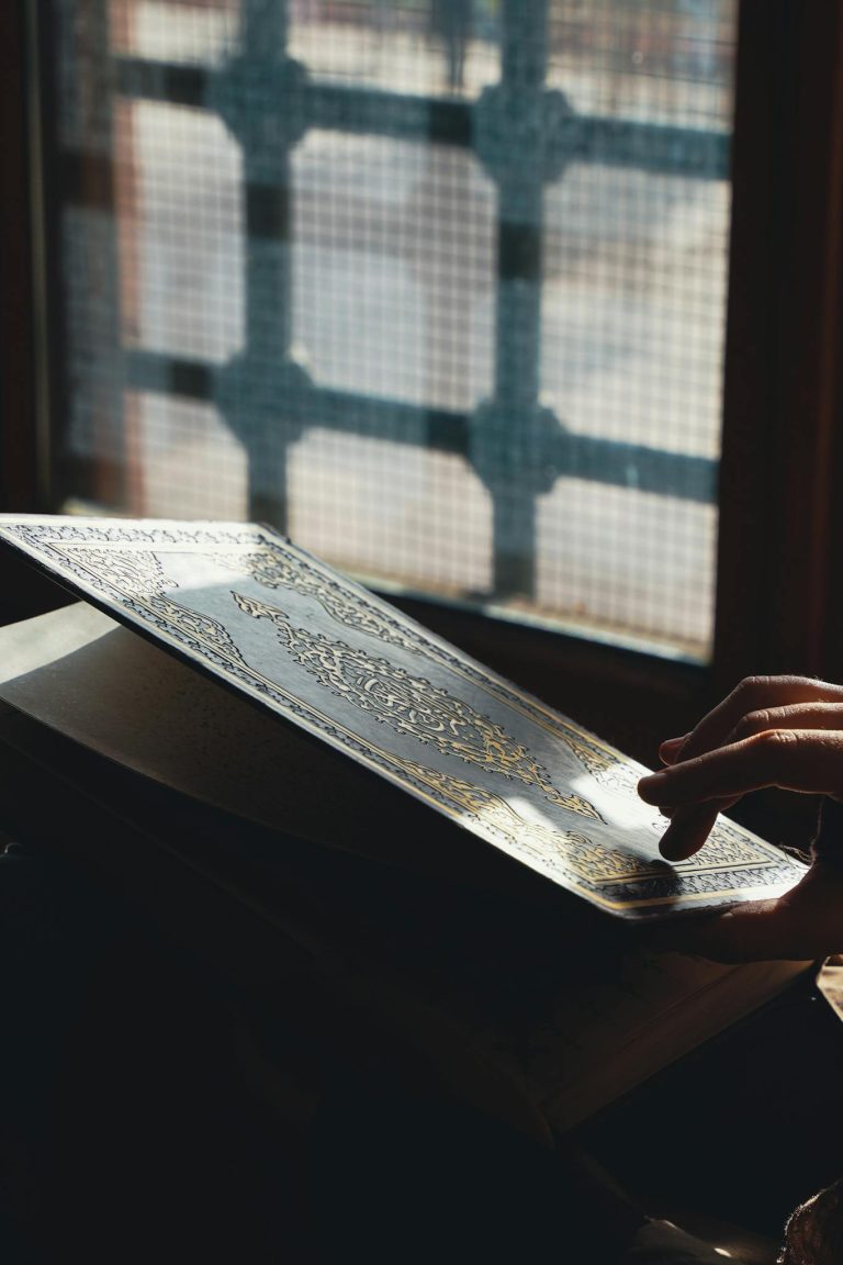 A close-up of a hand holding the Quran near a mosque window in Adana, Türkiye.
