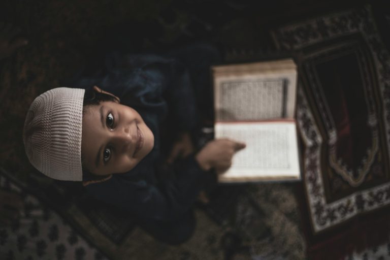 A smiling young boy in traditional attire reading the Quran on a prayer mat, viewed from above.